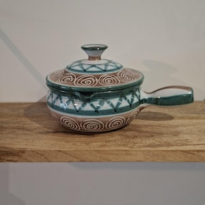 Peut inclure: Un plat de service en céramique avec couvercle et poignée. Le plat présente un motif en spirale et géométrique de couleur sarcelle, marron et blanc. La poignée et le bouton du couvercle sont également sarcelle et marron. Le plat est posé sur une surface en bois.