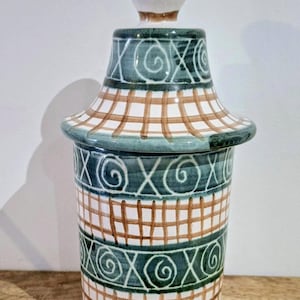 Peut inclure: Un pot en céramique décoratif avec un couvercle. Le pot présente un motif répété de carrés et de spirales turquoise, marron et blancs. Le couvercle a un petit bouton rond. Le pot est posé sur une surface en bois.