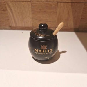 Pode incluir: Um pequeno pote de mostarda redondo de cerâmica preta com tampa a condizer e uma colher de madeira clara. A parte frontal apresenta um texto dourado que diz MAILLE DEPUIS 1747, acompanhado por um pequeno brasão e delicados motivos decorativos dourados.