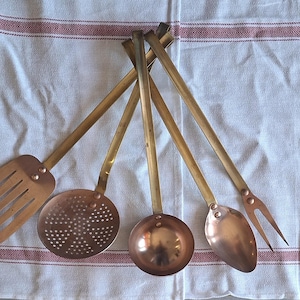 Peut inclure: Ensemble d'ustensiles de cuisine anciens en cuivre et laiton, comprenant une spatule ajourée, une écumoire, une louche, une cuillère et une fourchette. Les ustensiles ont des manches en laiton et des têtes en cuivre, sur un tissu blanc à rayures rouges.