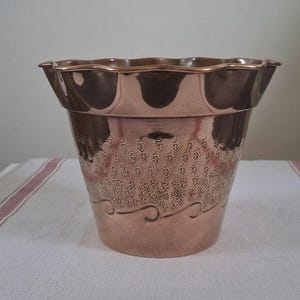 May include: A copper-coloured planter with a scalloped rim. The planter has a textured pattern on the body and a decorative wave design near the base. The planter is on a white surface.