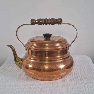 Peut inclure: Une théière en cuivre avec une finition martelée. Elle possède un bec et une anse en laiton, une poignée en bois et un bouton noir sur le couvercle. La théière est posée sur une surface blanche.
