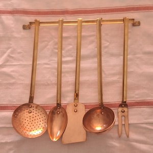 Könnte beinhalten: Ein Set aus fünf Küchenutensilien aus Messing, die an einer Messingstange hängen. Das Set umfasst einen Schöpflöffel, einen Löffel, einen Pfannenwender, eine Kelle und eine Gabel. Die Utensilien sind aus Messing und vor einem weiß-rot gestreiften Hintergrund angeordnet.