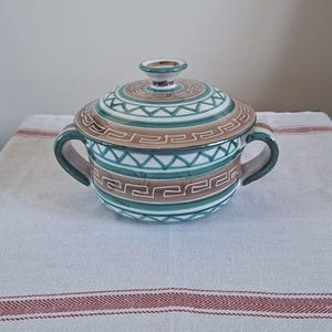 Peut inclure: Un bol en céramique avec couvercle, orné d'un motif géométrique turquoise, marron et blanc. Le bol possède deux anses et un petit bouton sur le couvercle. Le design présente des formes et des lignes répétitives.