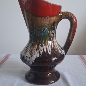 Peut inclure: Une cruche en céramique vintage au design distinctif. Le dessus est rouge brillant, passant à des motifs marron, verts et blancs. La base est marron foncé et comporte une poignée incurvée. Une pièce décorative pour la maison.