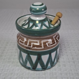 Peut inclure: Un pot en céramique avec un couvercle et une petite cuillère en bois. Le pot est décoré d'un motif géométrique dans des tons de vert et de marron. Le couvercle a un dessus arrondi avec un design assorti.