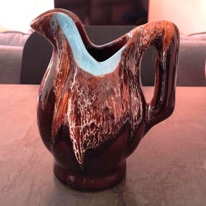 Peut inclure: Une cruche en céramique marron avec une finition brillante et un intérieur bleu clair. La cruche a une anse et un bec verseur. L'extérieur présente un motif unique de stries marron et blanches. La cruche est posée sur une surface grise.