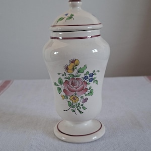 Può includere: Un vaso in ceramica bianca con coperchio, con un design floreale in rosa, giallo e blu. Il vaso ha una base a piedistallo e un bordo marrone. Il coperchio è sormontato da un pomello marrone. Un oggetto decorativo per la casa.