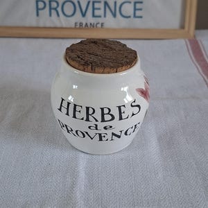 Puede incluir: Un tarro de cerámica blanca con cuerpo redondeado y tapa de corcho marrón texturizado. El frente del tarro presenta texto impreso en negro que dice HERBES de PROVENCE, acompañado de una pequeña ilustración floral en tonos rosa y verde.