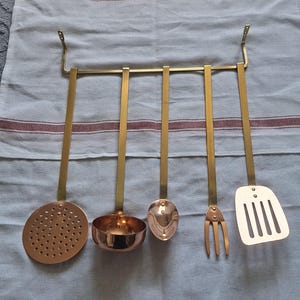Op de afbeelding: Een set van vijf messing keukengerei hangend aan een messing stang. De set bevat een schuimspaan, een pollepel, een lepel, een vork en een spatel. De keukengerei zijn gemaakt van messing en koper, tegen een lichtblauwe achtergrond met rode strepen.