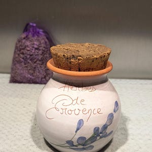 Peut inclure: Un pot en céramique avec un couvercle en liège, portant l'inscription "Herbes de Provence", orné d'accents floraux bleus. Un sac violet d'herbes séchées est visible en arrière-plan.