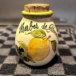 Peut inclure: Un pot en céramique avec un couvercle en liège, peint avec une poire jaune et des feuilles vertes. Le pot est étiqueté "Herbes de Provence" en écriture noire. Le pot a un bord vert.