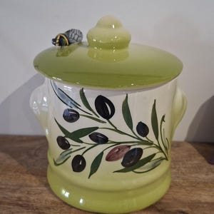 Peut inclure: Un pot en céramique avec un couvercle, peint avec des motifs de branches d'olivier. Le pot est principalement blanc avec une base et un couvercle vert clair. Une petite poignée en forme d'abeille est sur le dessus. Le pot est sur une surface en bois.