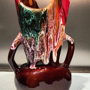 Peut inclure: Un vase en céramique vintage avec un design de glaçure unique. Le vase présente une base marron foncé, avec une section supérieure colorée dans les tons de rouge, vert, blanc et orange. Il a deux poignées et une large ouverture.