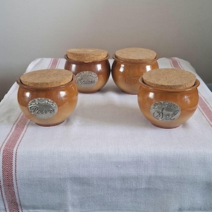 May include: Four ceramic spice jars with cork lids. The jars are a warm brown color with silver labels that read "Poivre", "Epices", and "Sel". They are arranged on a white tablecloth with red stripes.