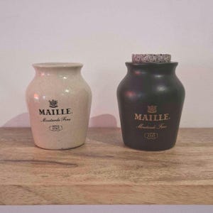 Può includere: Due barattoli di senape in ceramica, uno color crema e uno nero opaco, entrambi con la scritta nera MAILLE, Moutarde Fine e DEPUIS 1747. Il barattolo nero è chiuso da un tappo di sughero, mentre il barattolo color crema è aperto in alto.
