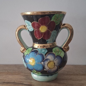 Könnte beinhalten: Eine schwarze Keramikvase mit zwei goldfarbenen Henkeln und einem goldfarbenen Rand. Die Vase ist mit handgemalten Blumenmotiven in Rot, Blau und Weiß mit gelben Zentren sowie grünen Blättern verziert. Der Vasenfuß ist in einem hellen Blauton gehalten.