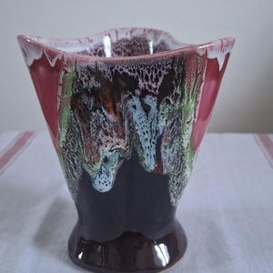 Peut inclure: Un vase en céramique vintage avec un glaçage texturé unique. Le vase présente un dessus carré avec un bord ondulé, se transformant en un corps évasé et une base arrondie. Les couleurs incluent le rouge foncé, le marron, le vert et le blanc, créant un aspect artistique saisissant.