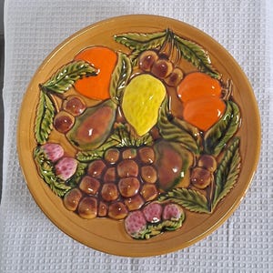 Peut inclure: Une assiette ronde en céramique avec un motif de fruits en relief. L'assiette présente des oranges, des cerises, des raisins, des fraises et un citron, le tout dans des couleurs vives. Des feuilles vertes entourent les fruits, sur fond beige.