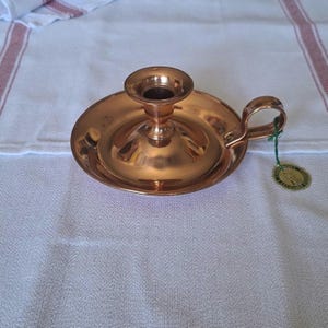 Peut inclure: Un bougeoir en cuivre avec une base ronde, une coupelle centrale pour la bougie et une poignée. Le bougeoir a une finition polie et une petite étiquette attachée à la poignée.