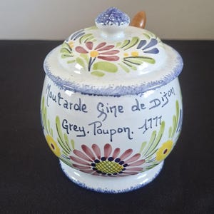 Op de afbeelding: Een witte keramische mosterdpot met deksel, versierd met kleurrijke bloempatronen en de tekst "Moutarde Fine de Dijon Grey. Poupon. 1777". Het deksel heeft een blauwe knop en een kleine houten lepel.