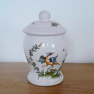 Peut inclure: Un pot en céramique blanc avec un couvercle, orné d'un motif peint à la main représentant un oiseau aux accents bleus, oranges et verts. Le pot a une forme arrondie et un petit bouton sur le couvercle. La végétation verte entoure l'oiseau.