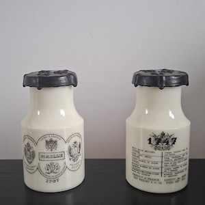 Może przedstawiać: Dwa kremowe ceramiczne słoiki z musztardą z czarnymi wieczkami. Słoiki mają etykiety z tekstem i herbami. Jeden słoik ma napis "MAILLE" i "1747".