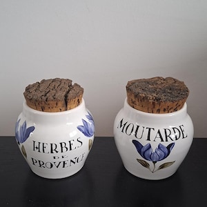 Peut inclure: Deux pots en céramique blanche avec des couvercles en liège. L'un porte l'inscription "HERBES DE PROVENCE" et l'autre "MOUTARDE" en écriture noire. Les deux pots présentent des motifs floraux bleus.