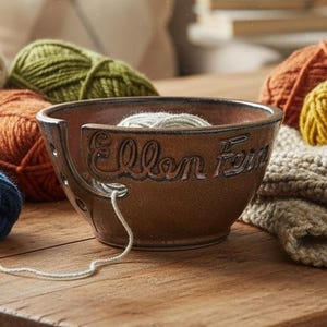 May include: A brown ceramic yarn bowl with the text Ellen Fern embossed on the side, featuring a spiral cutout for yarn. The bowl is surrounded by multiple skeins of yarn in cream, olive green, orange, mustard yellow, and blue, plus a knitted fabric.