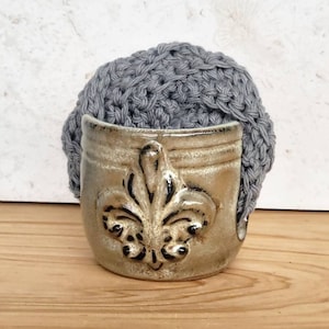 May include: A gray crocheted ball sits in a ceramic holder with a fleur-de-lis design. The holder is a light brown color with a darker brown glaze.