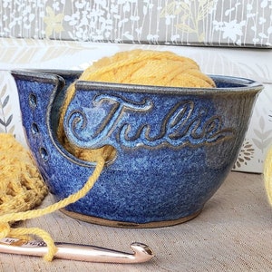 Blue yarn bowl with custom name Julie with ball of yellow yarn in it. Yarn is attached to a crochet project and hook.