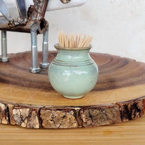 May include: A small, light green ceramic jar with a round body and a slightly flared lip. The jar is filled with wooden toothpicks.