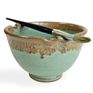 May include: A green ceramic bowl with brown glaze and a paintbrush resting on the rim.