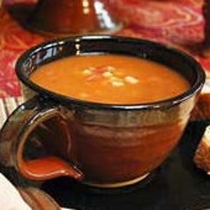 May include: A bowl of tomato soup with corn, beans, and other vegetables. The soup is served in a brown ceramic bowl with a handle.