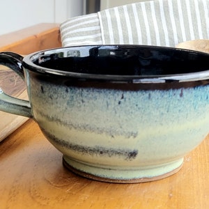 May include: A blue and white ceramic soup bowl with a black rim and a handle. The bowl has a speckled glaze and a slightly uneven finish.