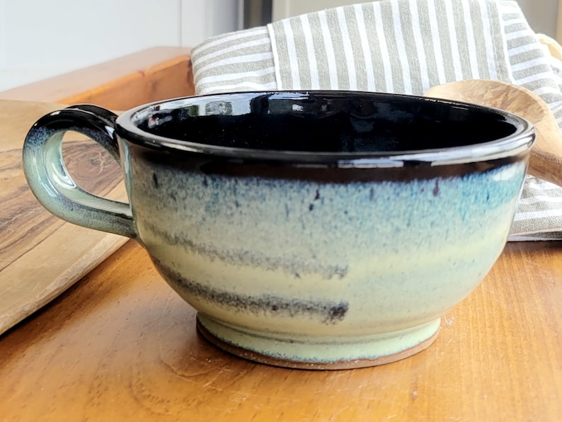 May include: A blue and white ceramic soup bowl with a black rim and a handle. The bowl has a speckled glaze and a slightly uneven finish.