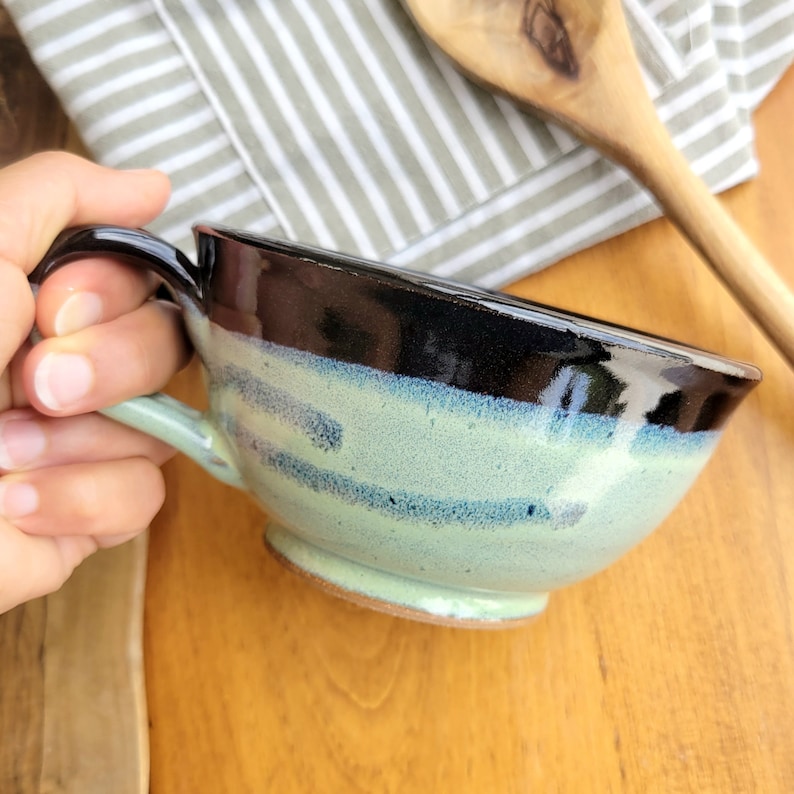 May include: A handmade ceramic bowl with a black rim and a light blue and green glaze. The bowl has a rounded shape and a handle.