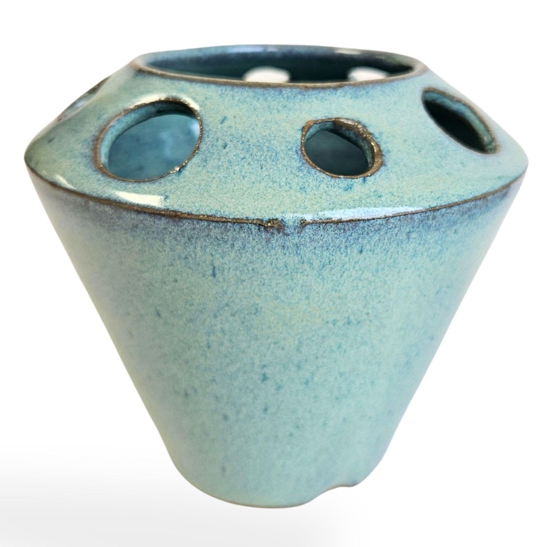May include: A light blue ceramic vase with six holes around the top rim. The vase has a speckled glaze and a rounded, flared base.