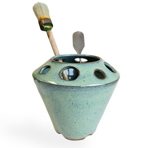May include: A light blue ceramic brush holder with a rounded base and multiple holes for holding brushes. A wooden handled brush and a metal spatula are in the holder.
