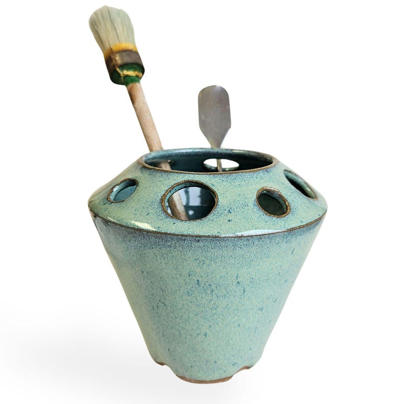 May include: A light blue ceramic brush holder with a rounded base and multiple holes for holding brushes. A wooden handled brush and a metal spatula are in the holder.