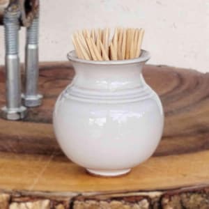 May include: A small, off-white ceramic vase with a rounded body and a narrow neck, filled with wooden toothpicks. The vase has horizontal ridges and sits on a wooden surface. The background includes metal bolts.