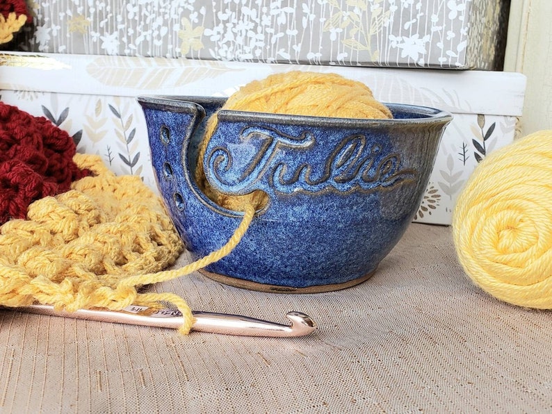 Blue yarn bowl with custom name Julie with ball of yellow yarn in it. Yarn is attached to a crochet project and hook.