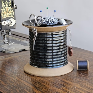 May include: A black ceramic sewing needle holder with a tan rim and a tan base. The holder has a decorative pattern and is filled with sewing supplies, including a pincushion, scissors, and a thimble. A spool of black thread sits on a wooden surface next to the holder.