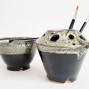 A set of 2 vessels intended for painters. The first is a black and white conical jar that tapers at the top and has compartment holes to store paint brushes. The other a pottery bowl having indentations in the rim for resting paint brushes.