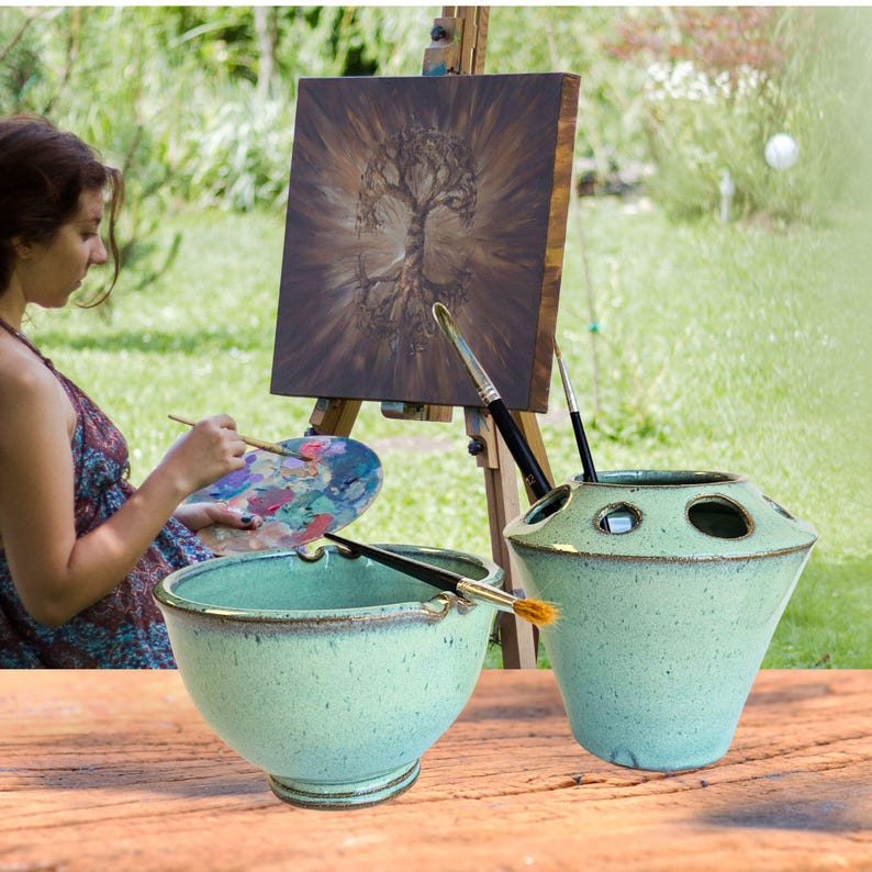 May include: Two ceramic bowls, one with a wide rim and the other with a narrow rim, both in a light green glaze with brown speckles. The bowls are used to hold paintbrushes and are sitting on a wooden table.