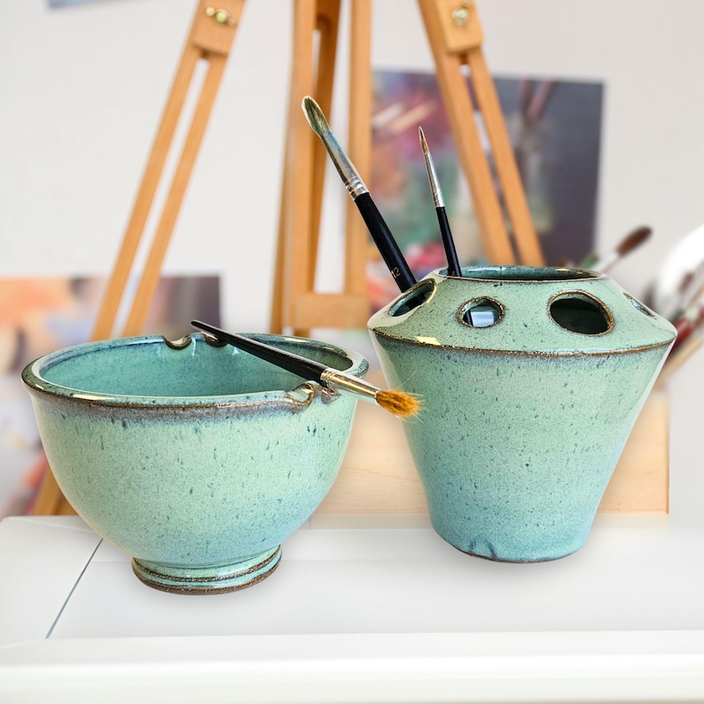 Artists painting set including paint brush holder and matching brush washing bowl with multiple storage slots and brush rests, holding paintbrushes in speckled green tones. The handmade wheel thrown pottery set is displayed in an artist’s studio with an easel and canvases and blurred artwork in the background.