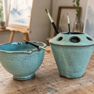 May include: A turquoise ceramic bowl with two notches on the rim and a matching turquoise ceramic brush holder with multiple circular openings, both featuring a speckled glaze finish and dark brown edges, holding several paintbrushes.