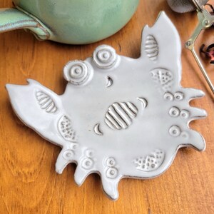 May include: A white ceramic crab-shaped teacup coaster with a textured surface and raised details. The coaster has a whimsical design with eyes, a mouth, and other decorative elements.