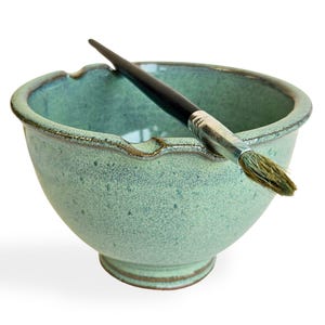 May include: A green ceramic bowl with a brush resting on the rim. The bowl has a speckled glaze and a slightly wavy edge.