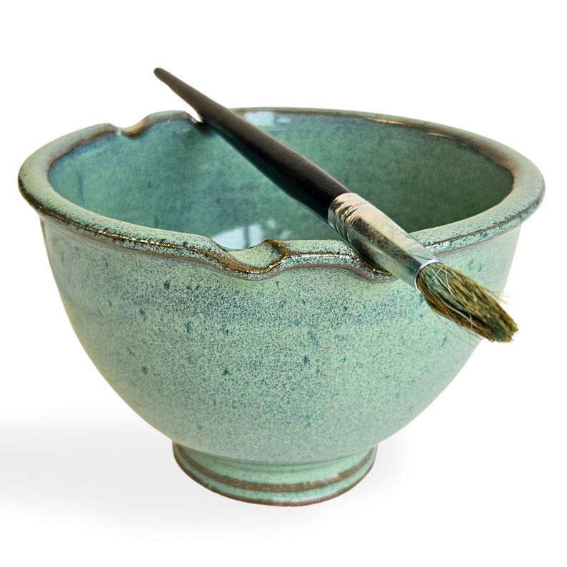 May include: A green ceramic bowl with a brush resting on the rim. The bowl has a speckled glaze and a slightly wavy edge.